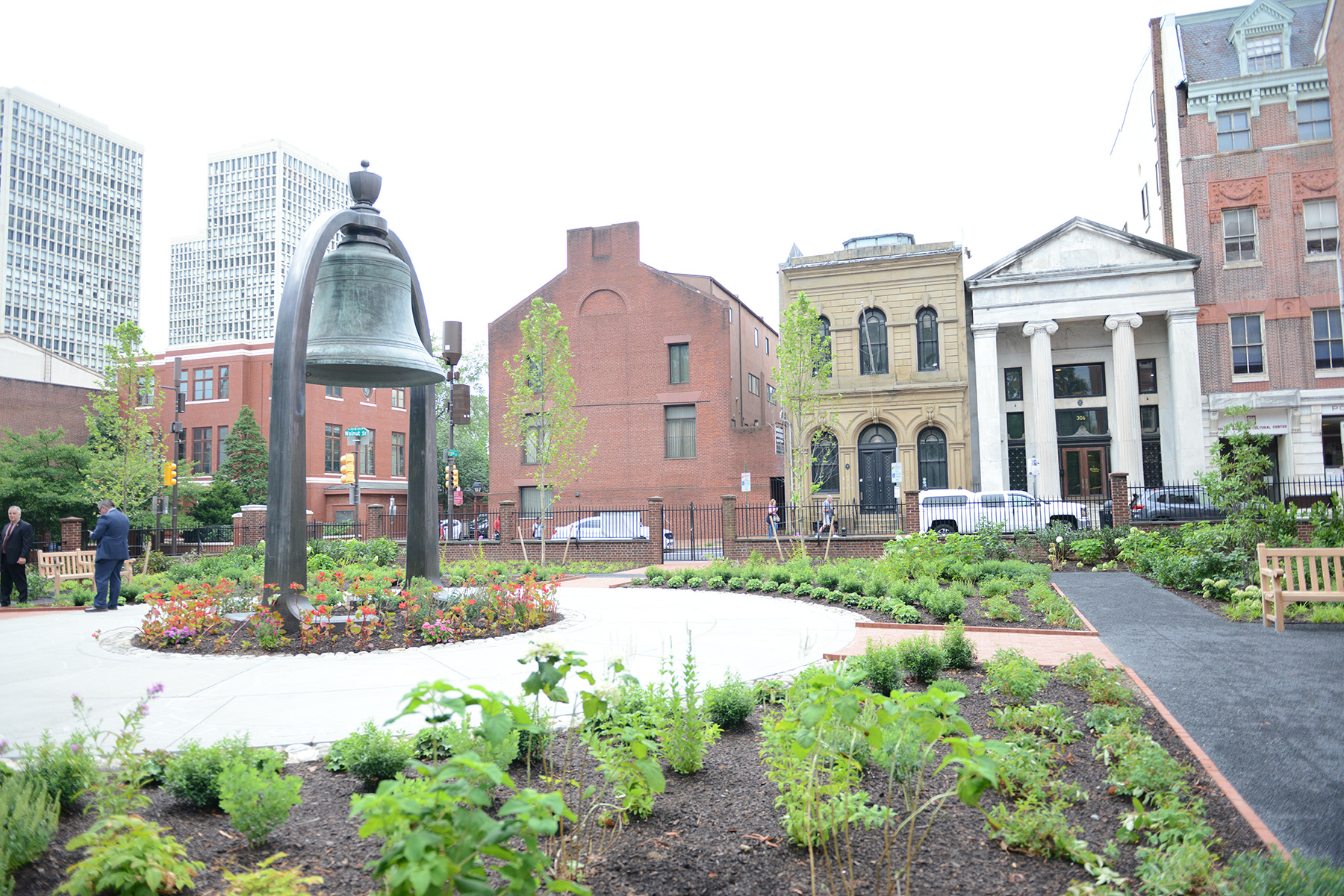 The bell is installed in its arch, with pathways, flowers, and green shrubs surrounding it. 
