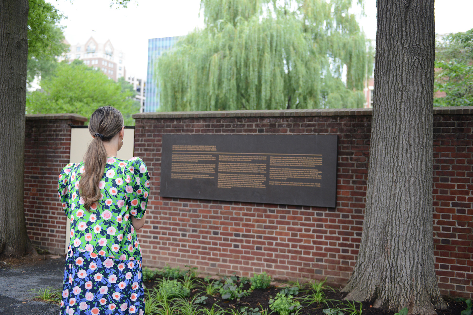 A woman with a ponytail, wearing a flowery dress, stands in front of signs installed on a brick wall. The black sign with gold letters explains the history of the bell and Benjamin Rush.