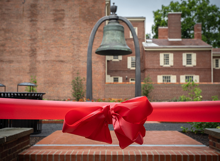 A blurred image of a six-ton bronze bell that hangs in a park, surrounded by brick buildings. A red bow is added over the image. 