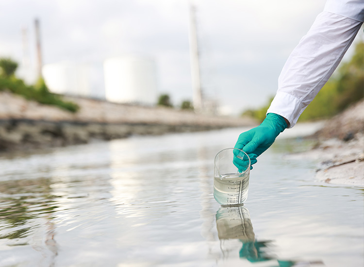 A green gloved hand for someone wearing a white protective suit takes a water sample using a clear beaker. 