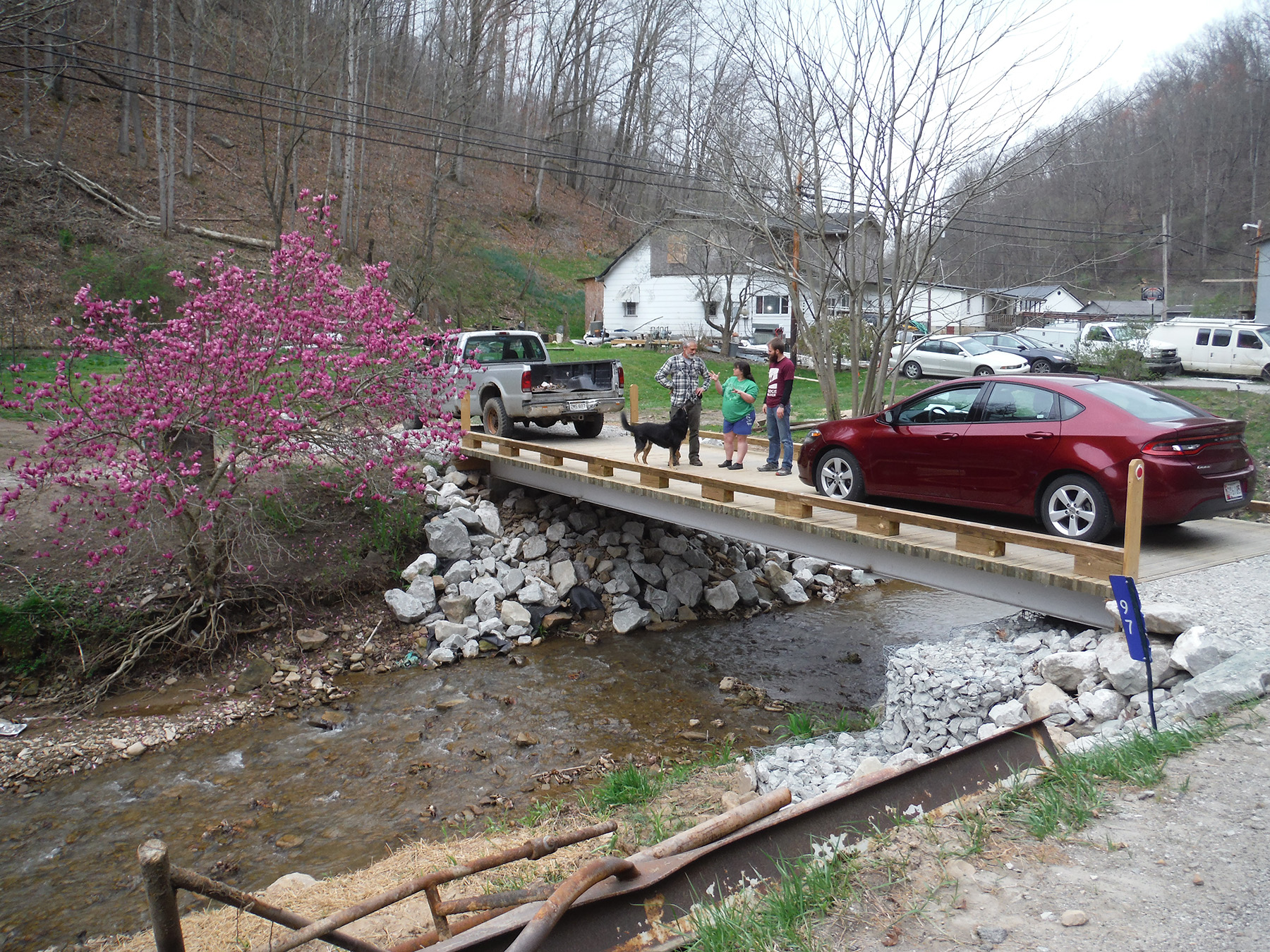 A maroon car and a silver truck are parked on a bridge. There are three people talking and a dog is also on the bridge. Below the bridge is water and in the background there is a flowering tree, bare trees, and a couple of houses. 