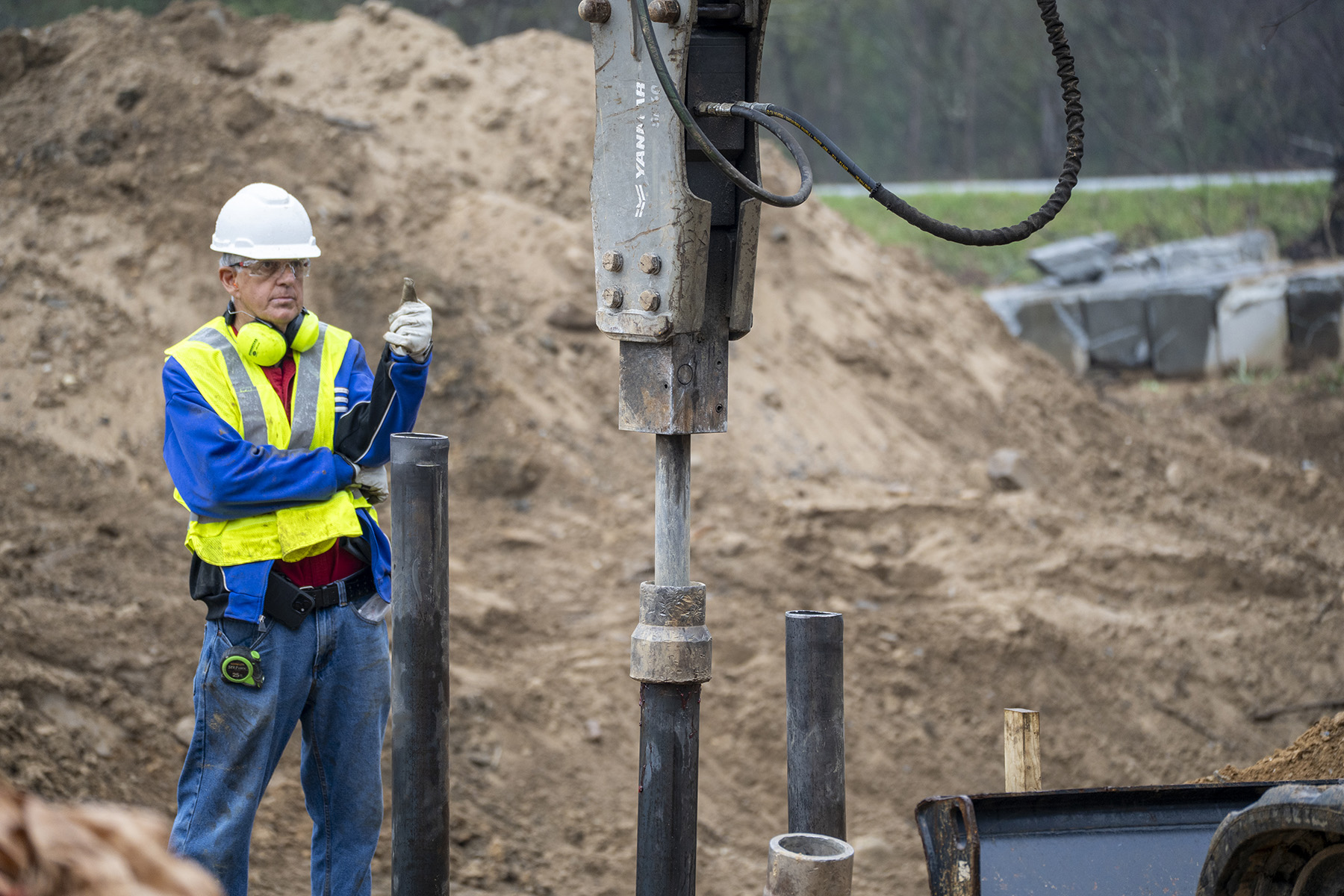A man in a yellow safety vest with headphones and a hardhat looks at machinery. 