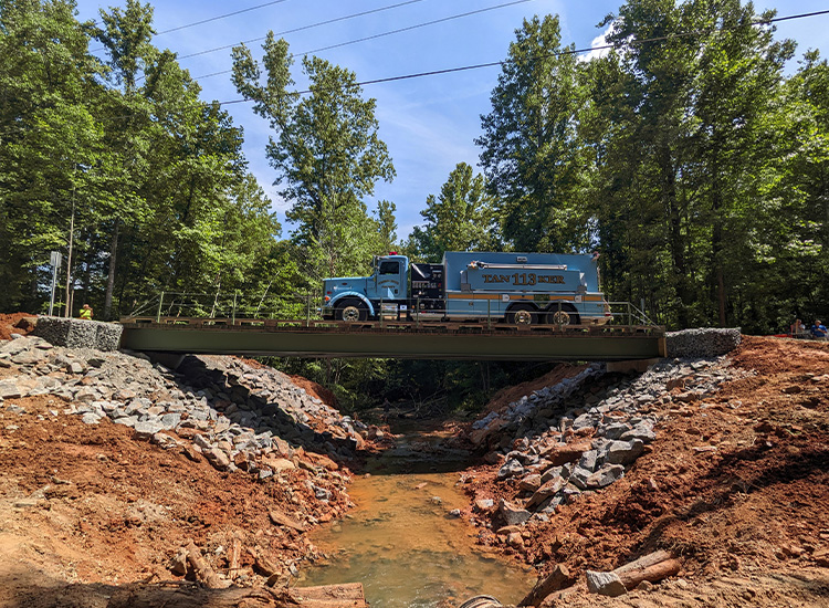 A blue commercial truck crosses a steel bridge. Below the bridge is water. 