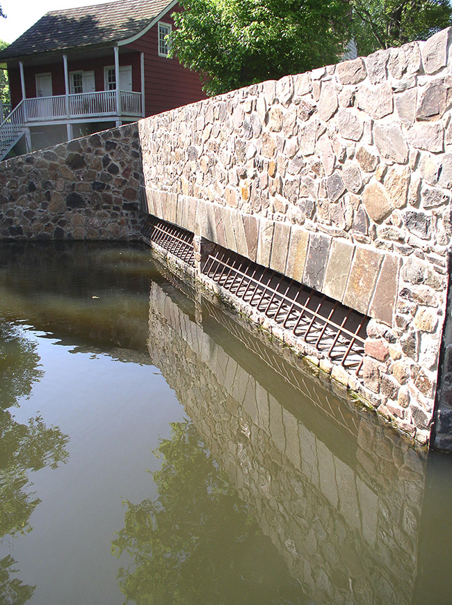 Stonework covers the face of a stormwater structure near a historic mill and pond. 