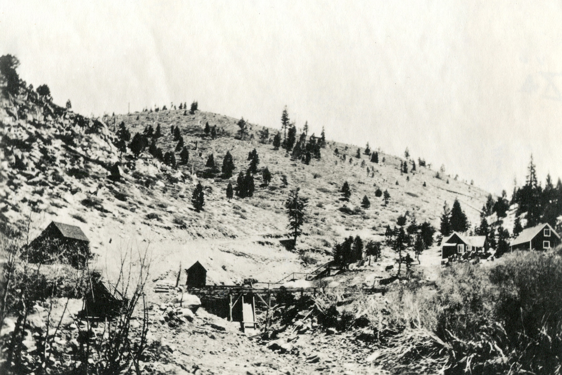 Black and white photo shows a hillside sparsely dotted with trees. IN the foreground are several cabins and the beginnings of a flume structure that would bring water down the hillside. 