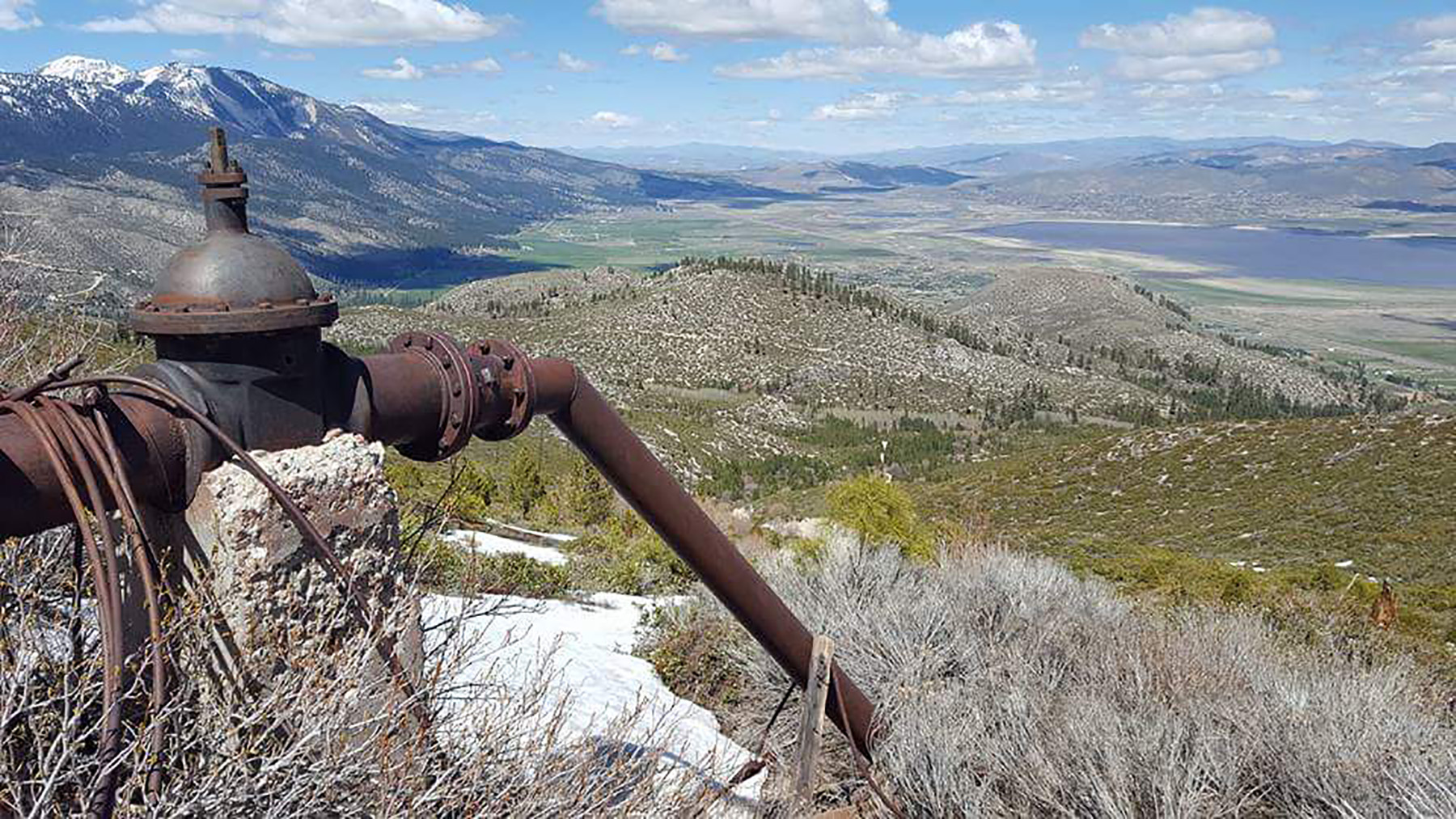 Image shows a rusty iron water valve and pipe leading down a hillside. 