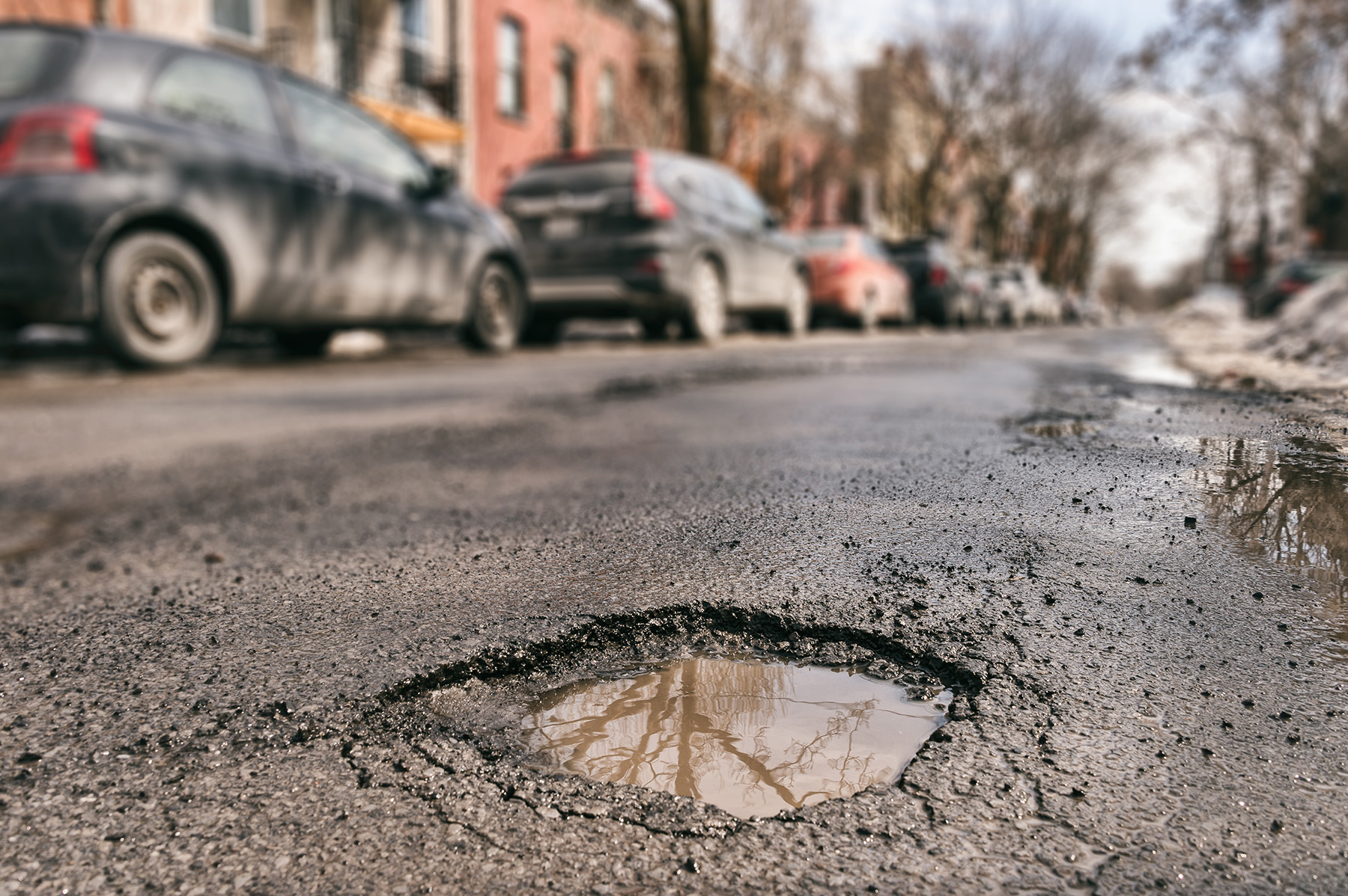 An up-close view of a small, rain-filled pothole on a street. In the blurred background is a line of cars parked on a curb. 