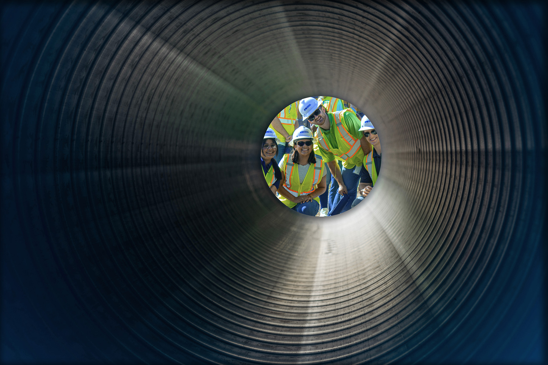 Four students wearing yellow safety vests, black sunglasses, and white hard hats smile into a ridged tunnel.