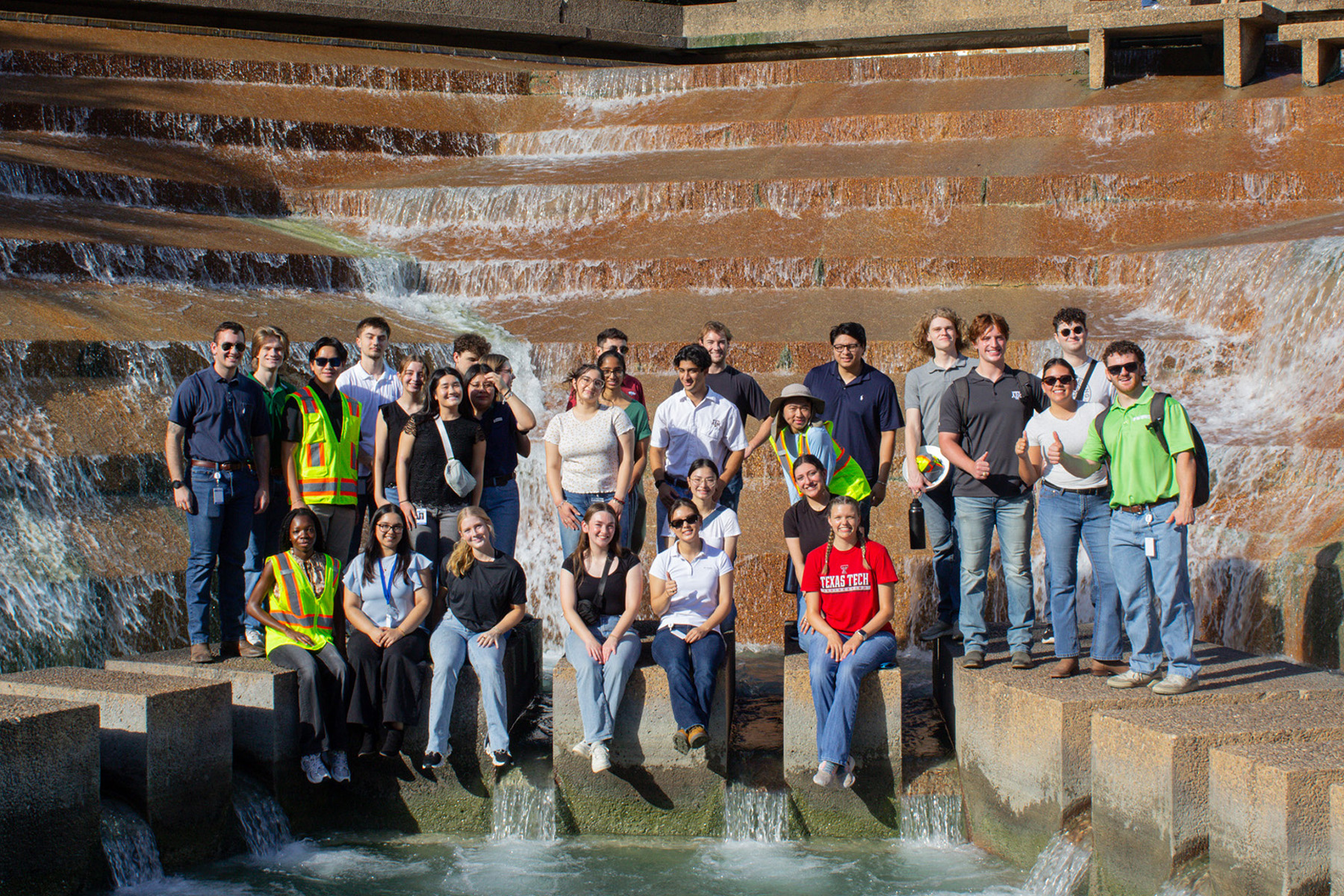 Twenty-nine people stand or sit in front of a waterfall for a group picture. Water pours down a rust-color stone wall behind them.