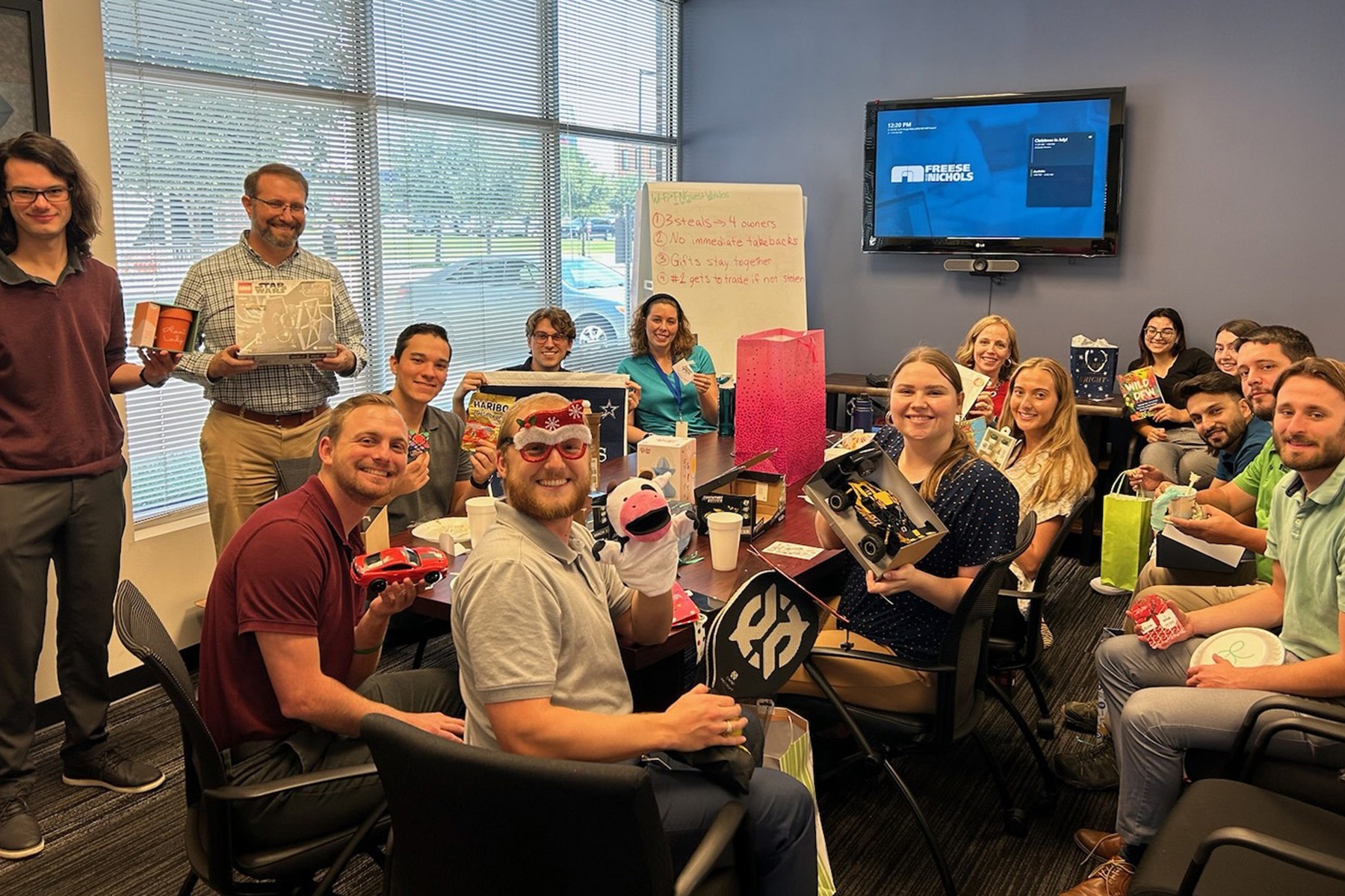A group of people sit around a table in an office meeting room showing small items to the camera. 