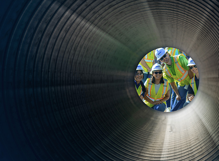 Four students wearing yellow safety vests, black sunglasses, and white hard hats smile into a ridged tunnel.