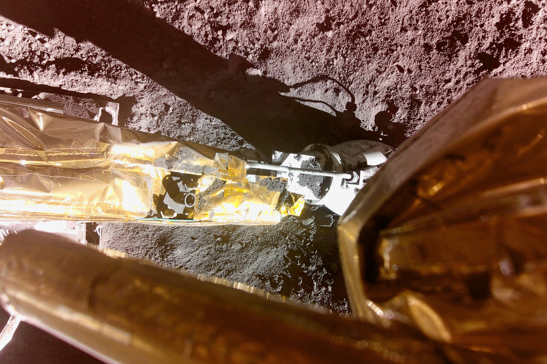 A picture taken of the moon's surface from inside the PlanetVac. A shiny gold leg and the white vacuum are visible, along with a shadow of the vacuum arm.