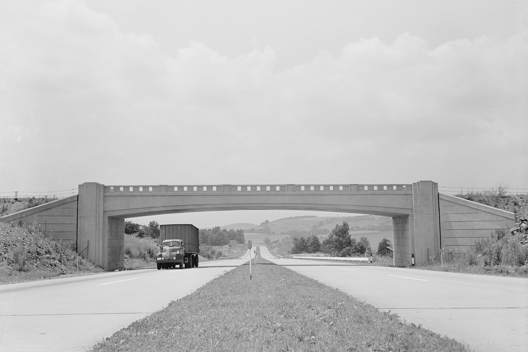 A truck drives on one side of a two-lane roadway. There is a grassy median between lanes and a bridge spanning the roadway. 
