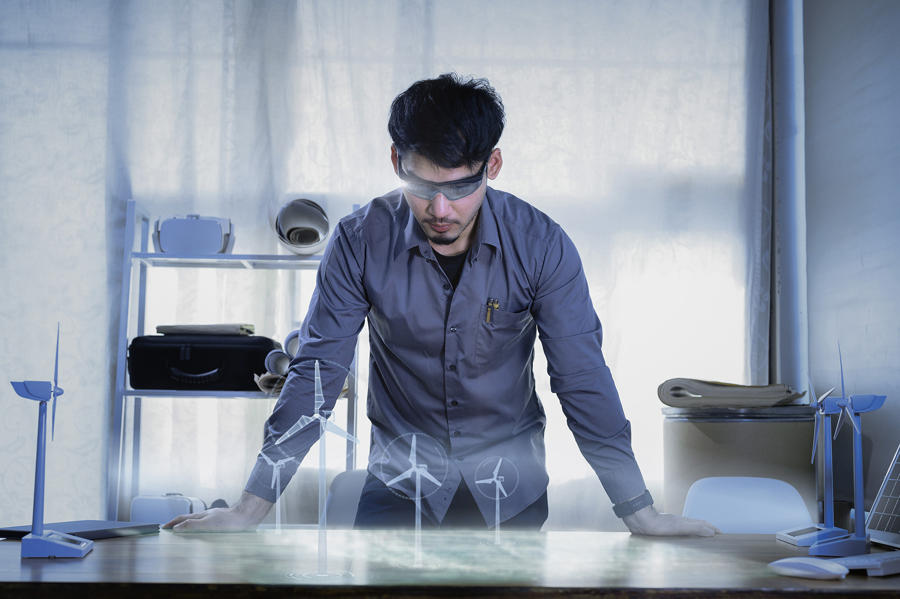 A man with black hair leans his hands on a desk, looking at a grouping of four virtually created 3-dimensional windmills. 
