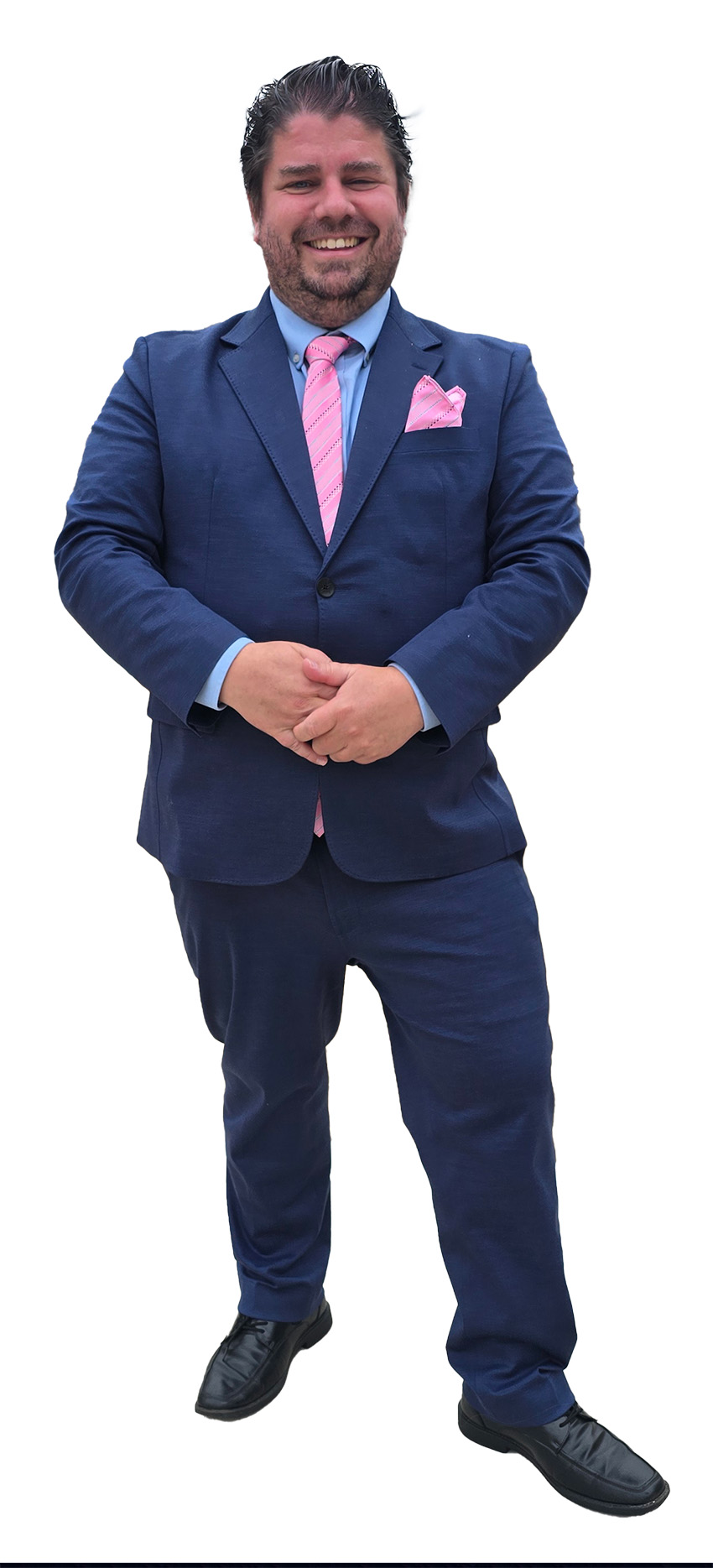 A man in a blue suit, blue shirt, and pink tie, his hands clasped before him, smiles at the camera. 
