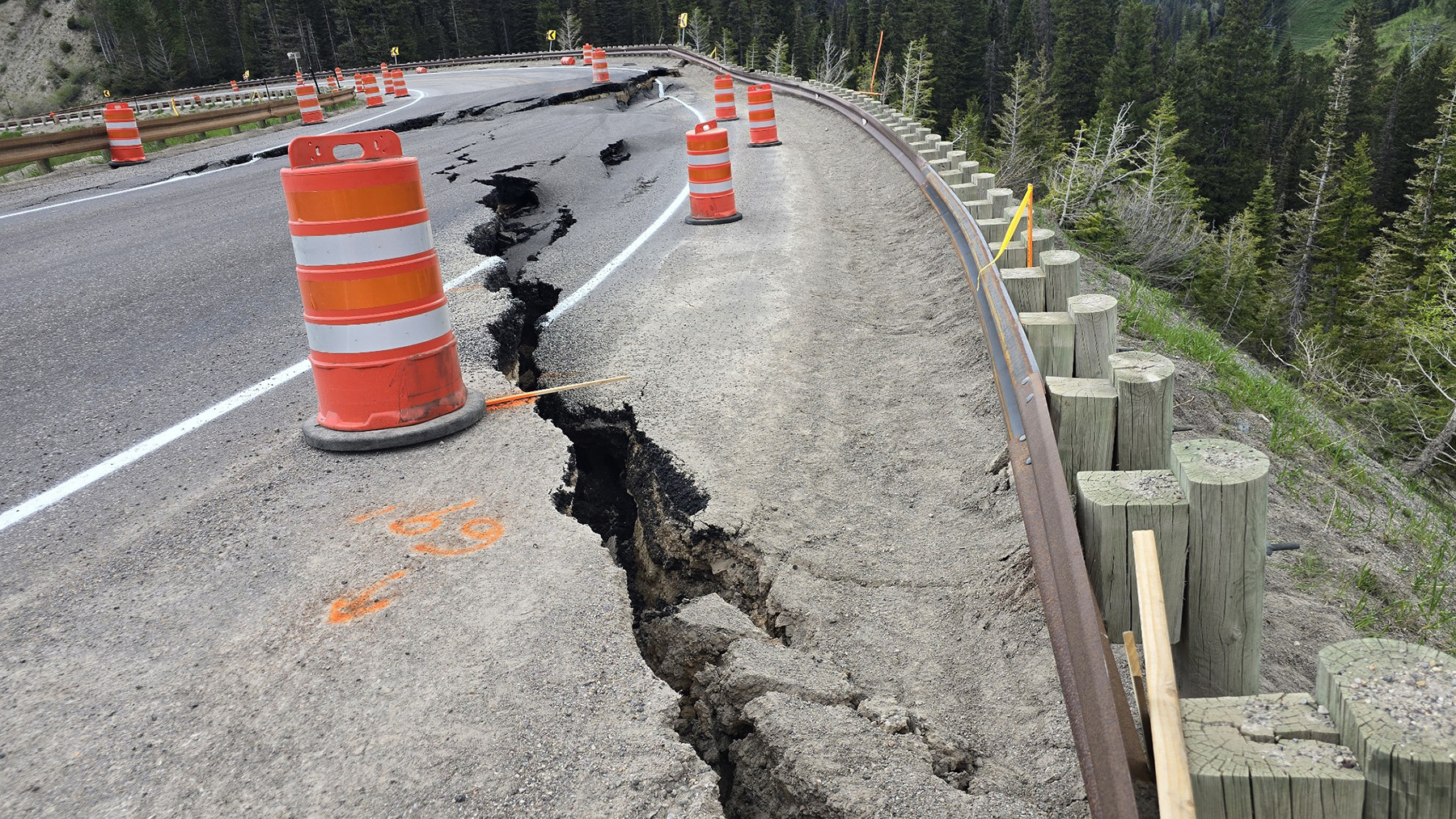 Pavement has a massive crack in it that stretches across the entire roadway. Orange and white construction barriers are on the roadway. 