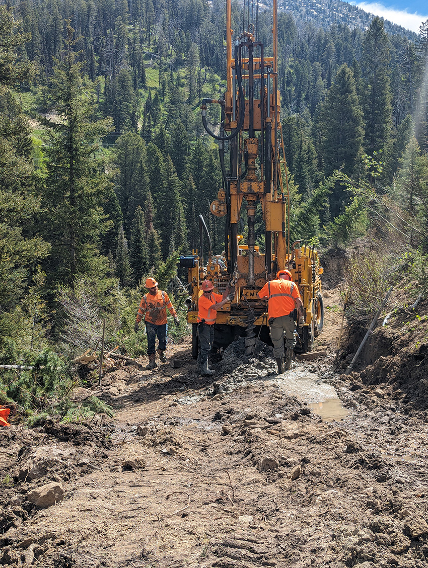 Men in orange shirts and vests wearing safety hats walk around in a muddy area drilling boreholes into the soil. 