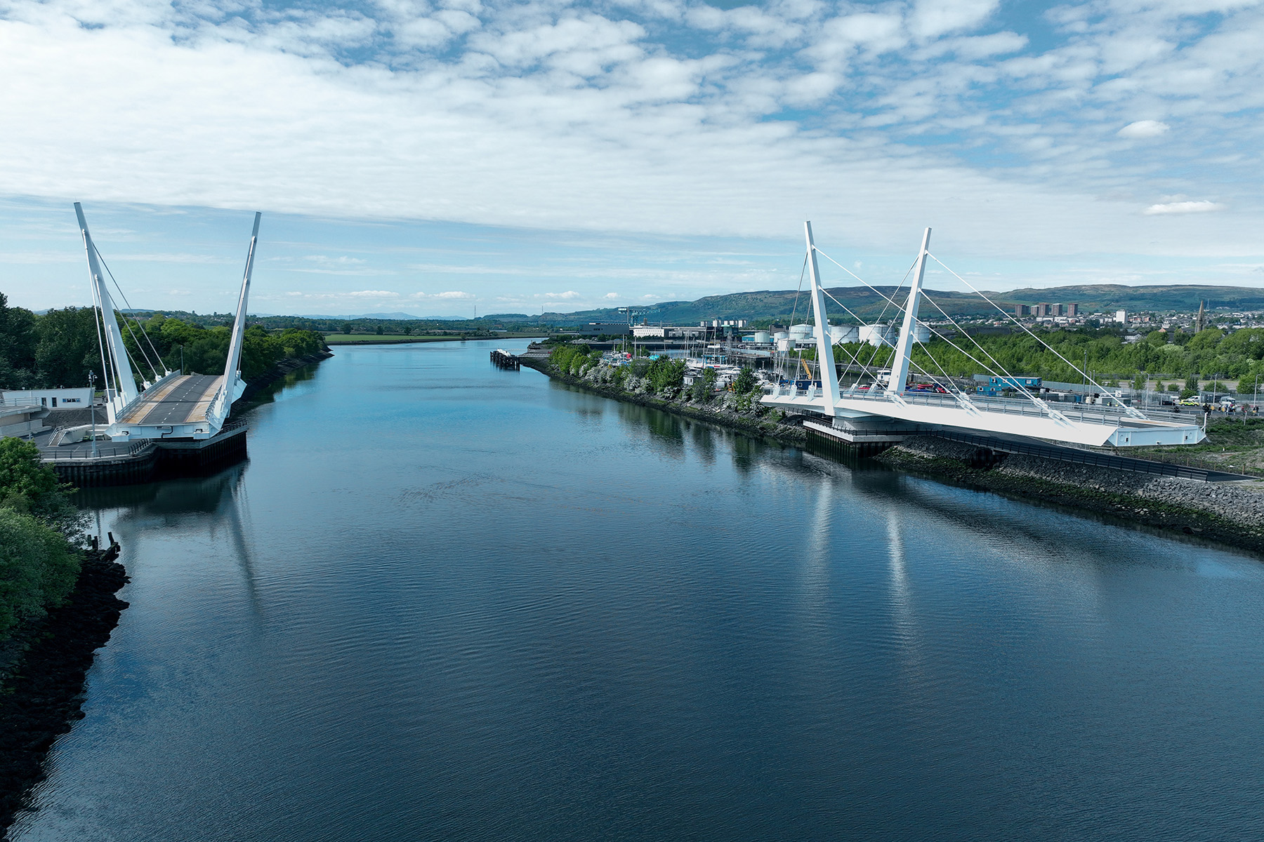 The two leaves of a swing bridge are open. The bridge traverses water. There is greenery on the right shore and a town and construction site on the left. 