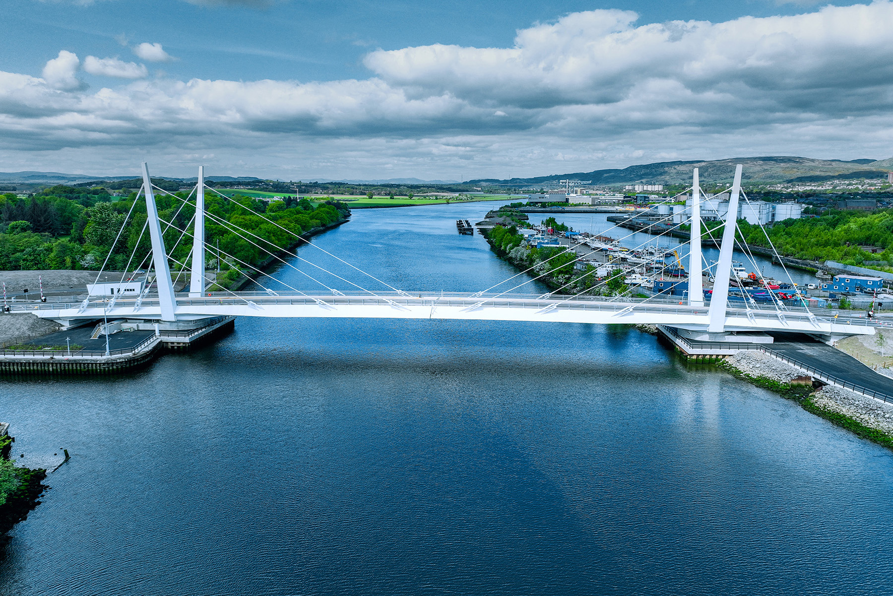 A white bridge spans a body of water. There are cables coming from the bridge deck and connecting to tall pylons. There are trees and grass in the foreground and background.