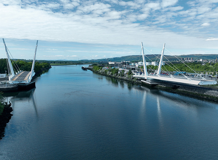 The two leaves of a swing bridge are open. The bridge traverses water. There is greenery on the right shore and a town and construction site on the left. 
