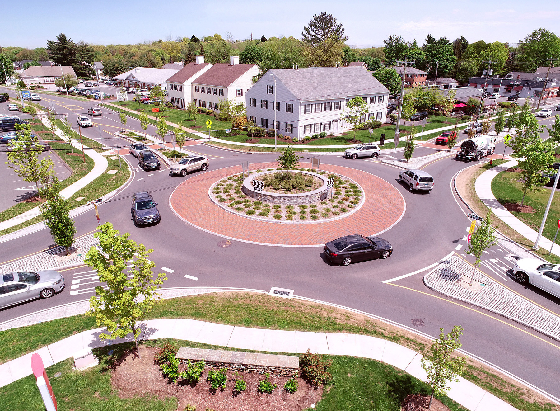 The photograph shows an aerial view of cars and other vehicles moving around a roundabout on a wooded street surrounded by residential and commercial buildings. 