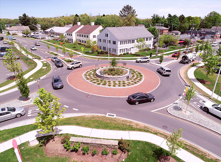 The photograph shows an aerial view of cars and other vehicles moving around a roundabout on a wooded street surrounded by residential and commercial buildings. 