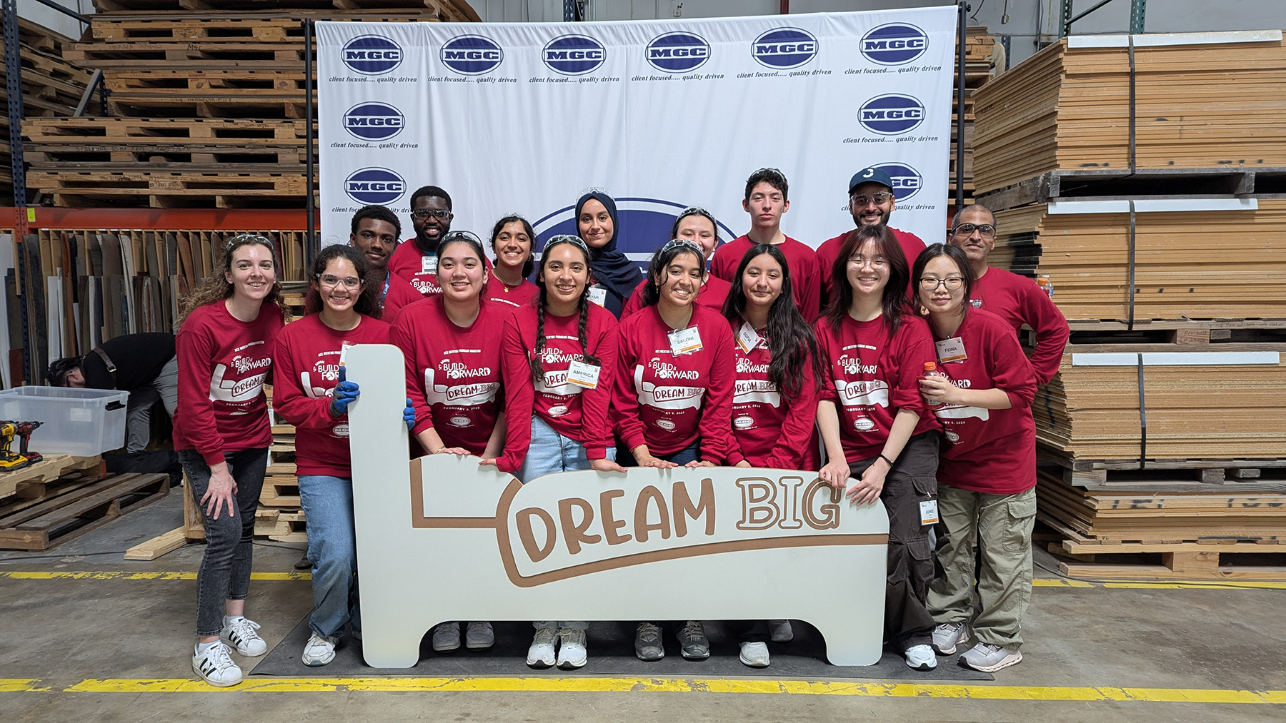 A group of young men and women, in matching red long-sleeve t-shirts stand behind a cardboard cutout of a bed bearing the phrase: Dream Big. 