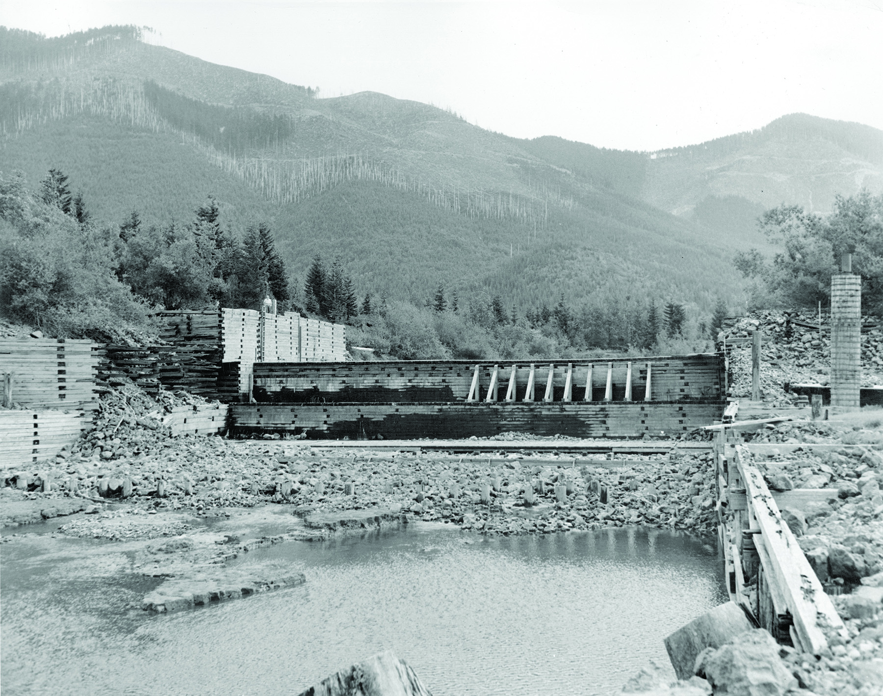 Image shows a wooden dam during construction. There is a low level of water in the foreground and on the sides of the dam is construction rubble. 