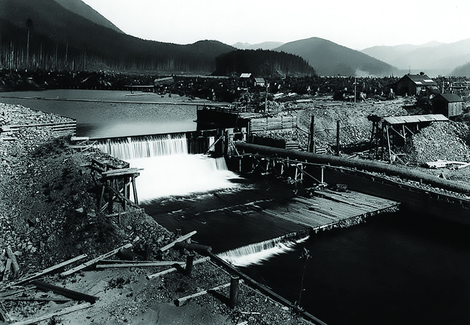 Image shows a dam with water running over the crest. There are wooden sheds and other construction paraphernalia in the background. 