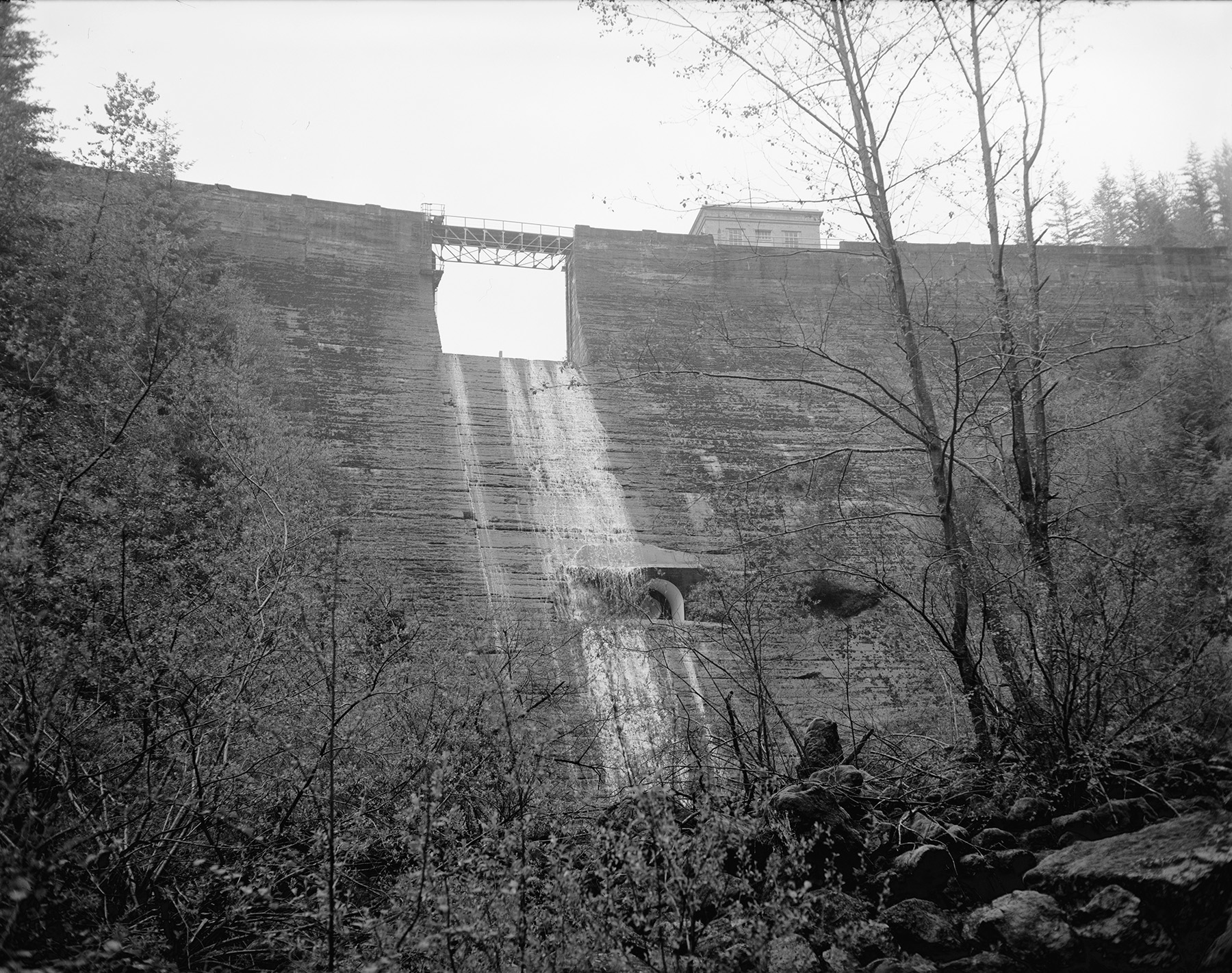A tall masonry dam can be seen. There is a spillway in the center of the dam. 
