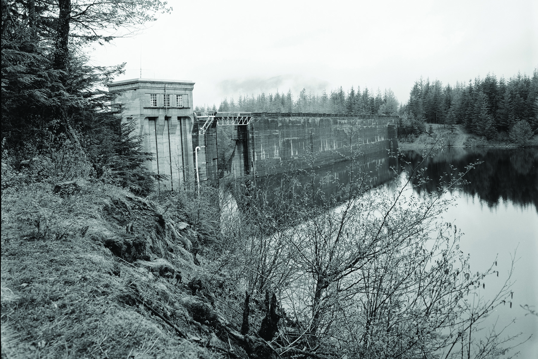 A masonry dam and its reservoir are shown in the photo. There are trees on either side of the dam. 