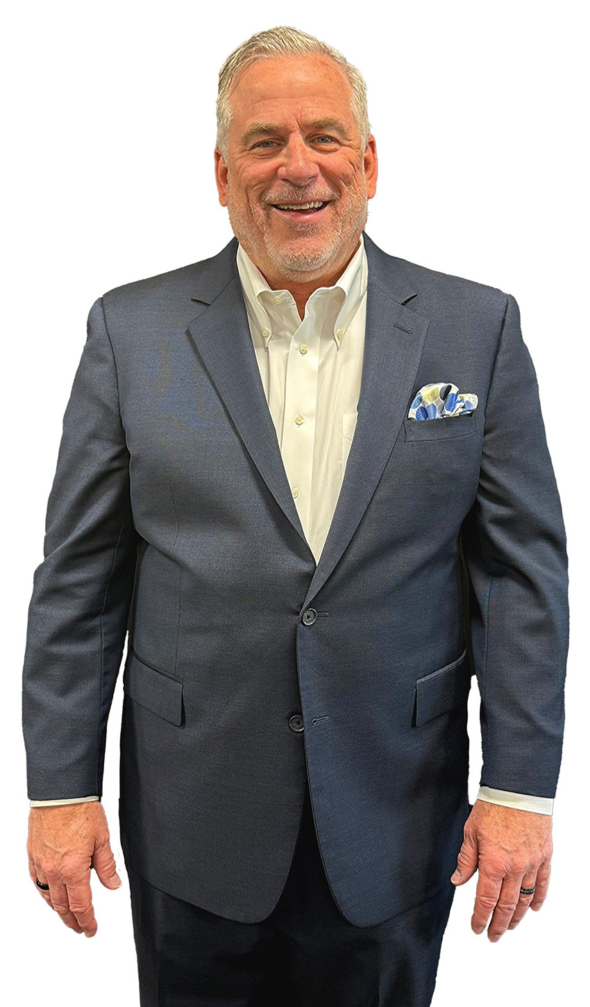 Man with graying hair and beard smiles at the camera. He’s wearing a button-up shirt and a gray suit. 