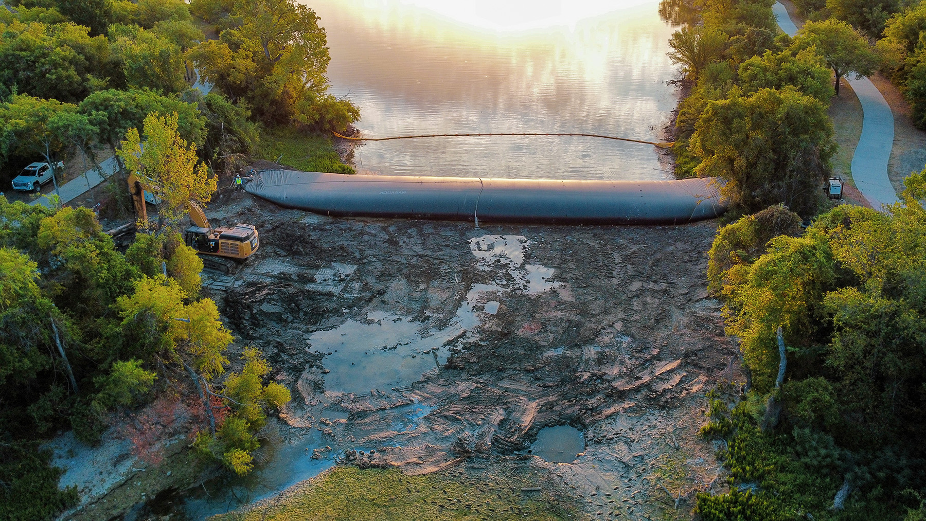A bird’s eye view of the lake shows a massive, inflated bladder dam in the middle. One side of the lake is dry and the other side is normal. There are trees on either side of the lake. 