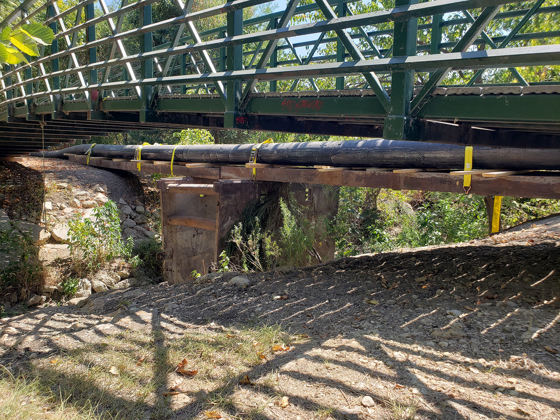 Next to a green pedestrian bridge is black piping elevated off the ground with wood slats attached to metal beams. The wood and piping are secured together with large yellow tie-downs.