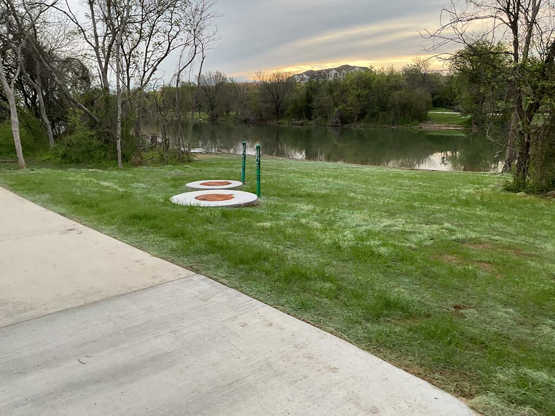 Two concrete circles with bronze metal manhole covers are in a grassy area. In the foreground is a sidewalk and in the background is a lake and trees. The trees are reflected in the still water.