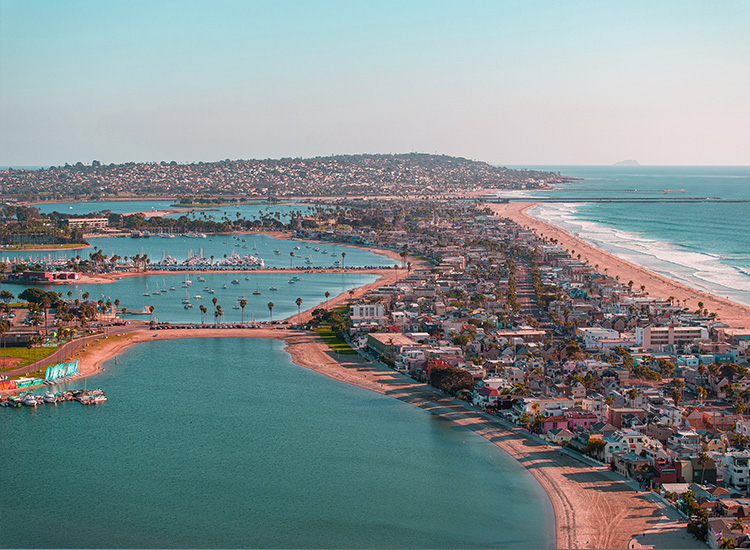 An aerial photograph shows coastal San Diego with a multitude of homes, commercial buildings, boats, as well as beachfront and water. 
