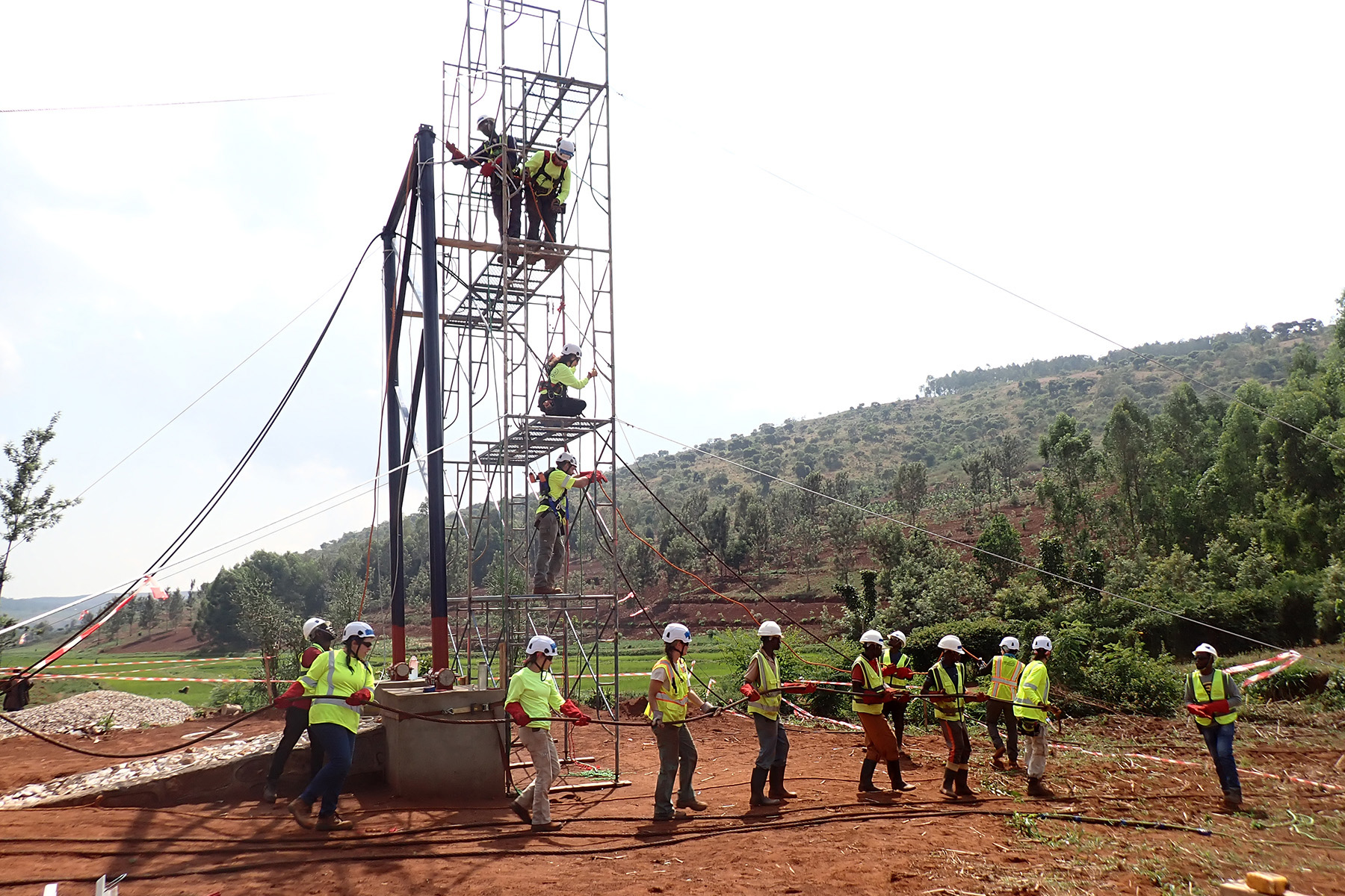 Volunteers in yellow jackets and white hard hats work on scaffolding on reddish dirt against a verdant background.