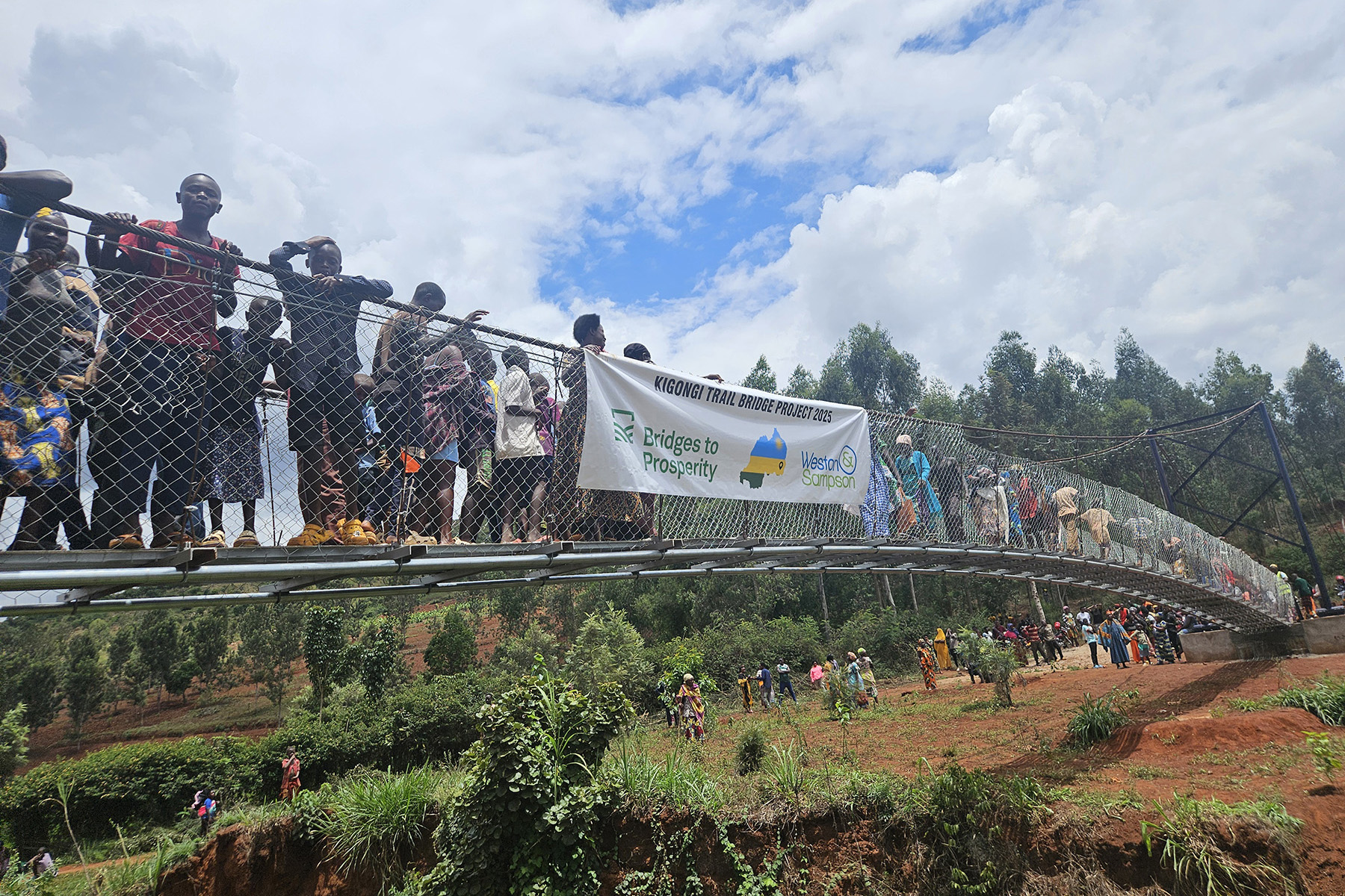 Dozens of people stand on the newly completed bridge, or in the background, with a cloth sign recognizing the volunteers.