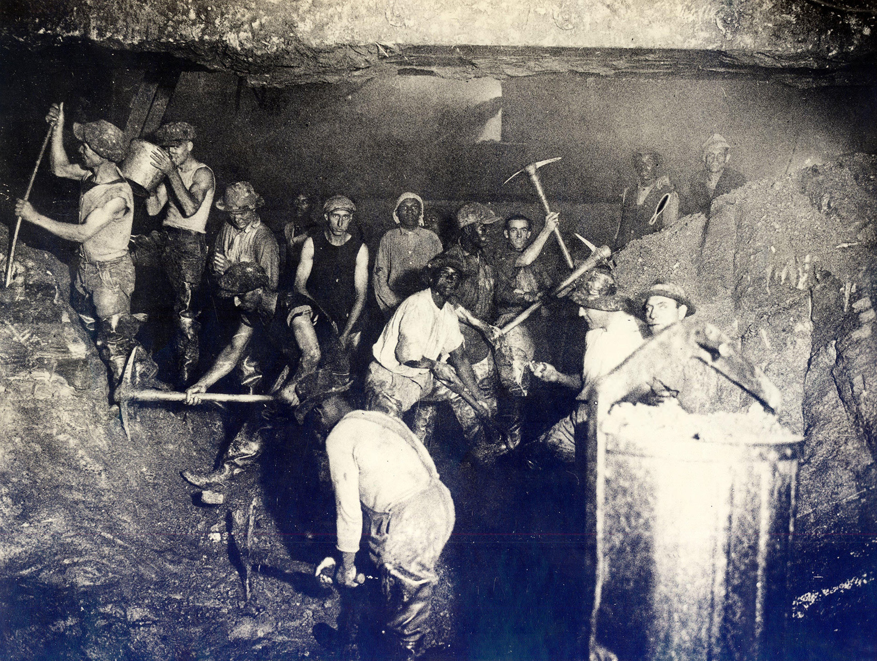 Men, some holding pickaxes or buckets, cut away at bedrock inside a caisson.