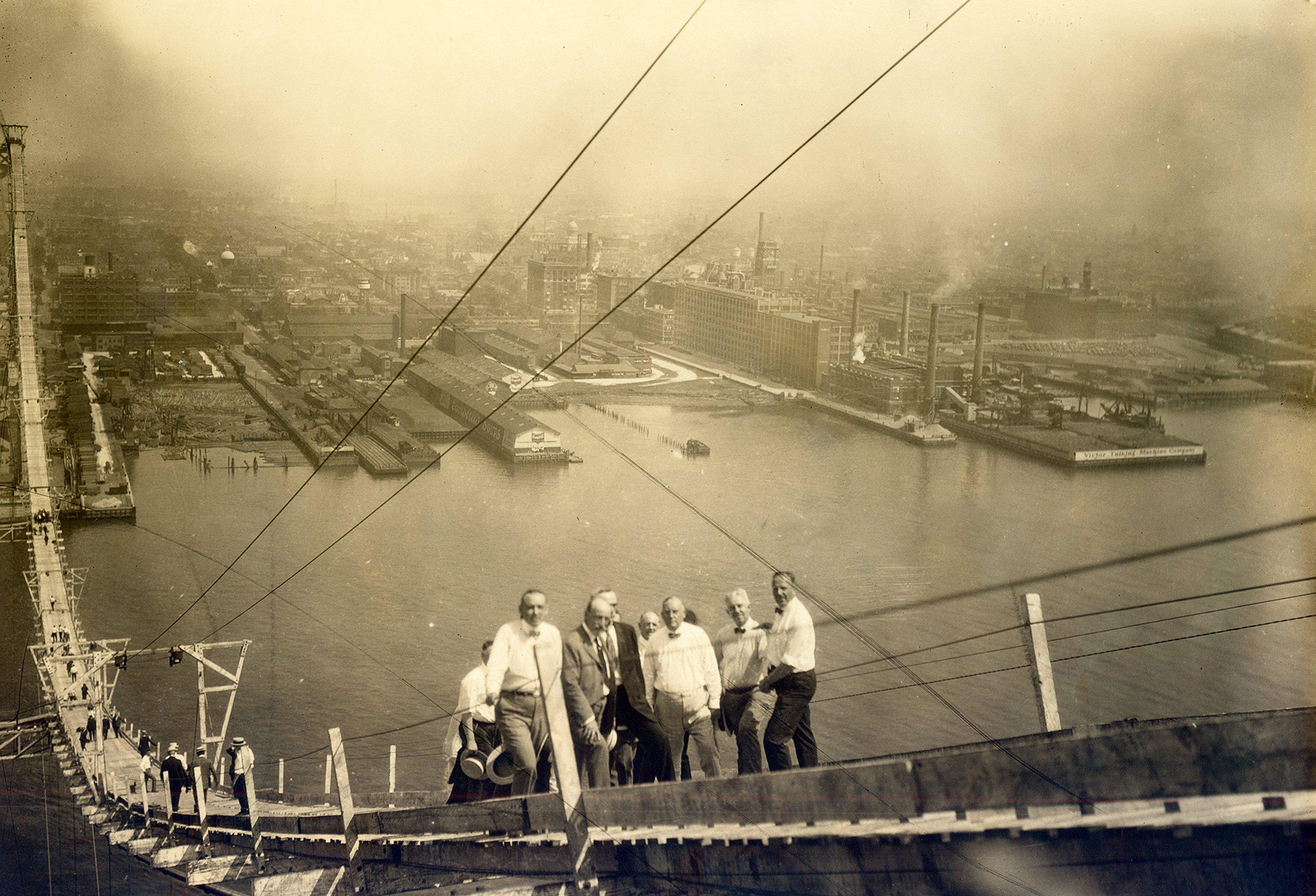 Photo shows a group of men walking across a wooden footbridge that spans a large body of water.