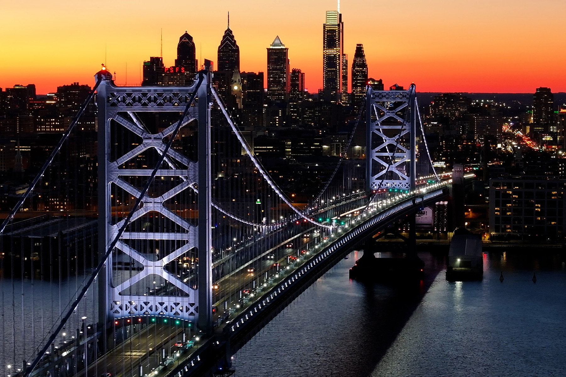 Picture shows a steel bridge lit up at sunset. Cars are driving on the bridge, and the cityscape can be seen in the distance.