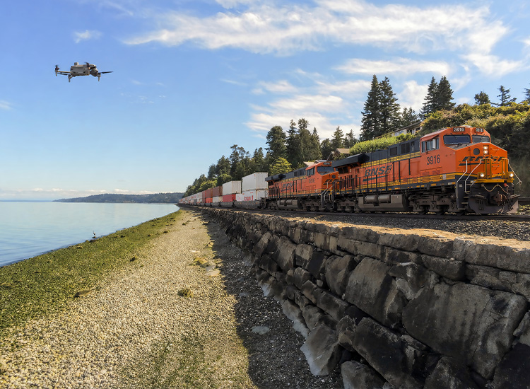 A panoramic view shows a drone flying alongside an orange BNSF freight train running along coastal tracks. 