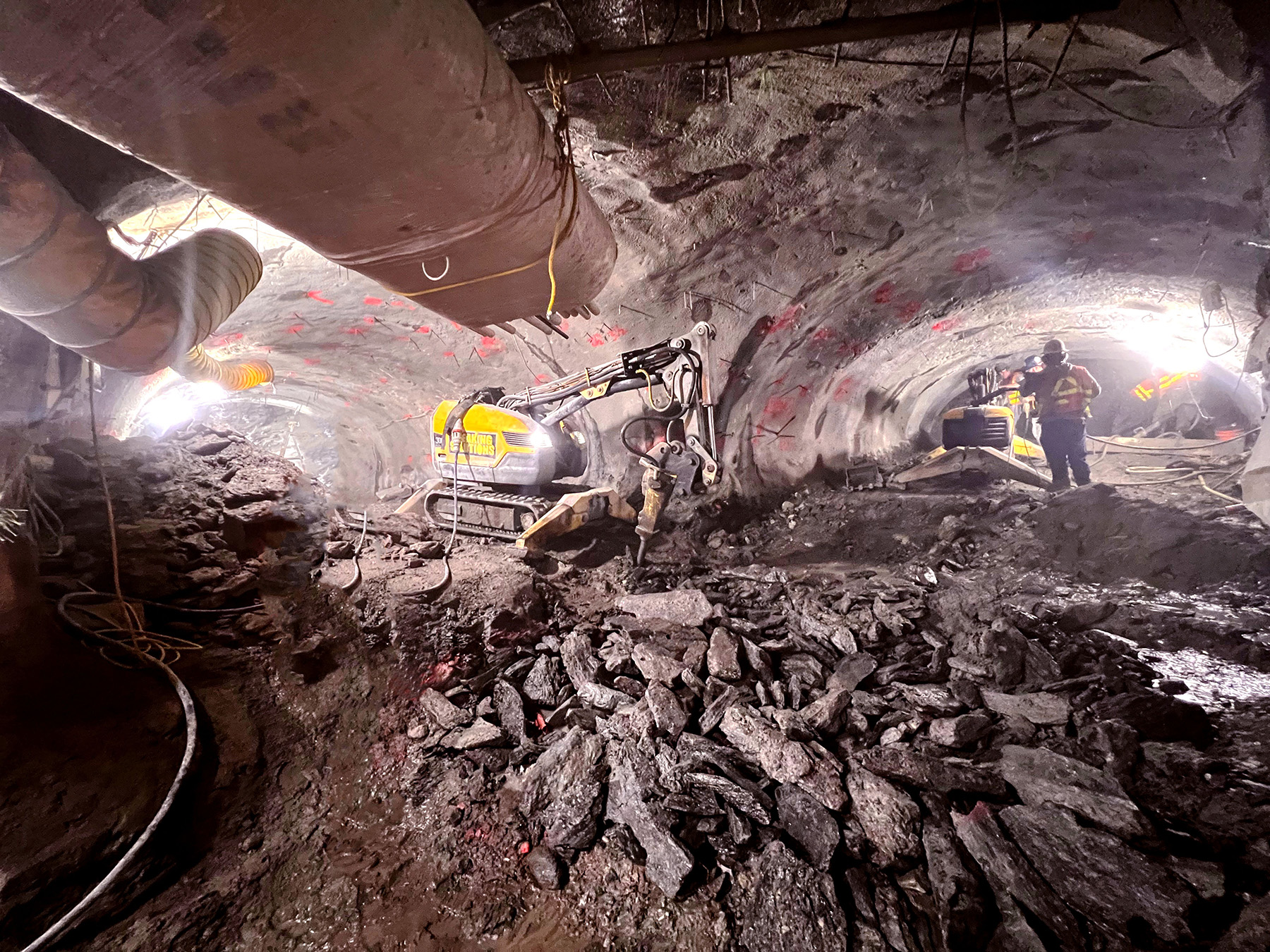 The tunnel under excavation. Piles of rock sit in the foreground. In the background are two yellow excavator, one on each side of the tunnel curve.