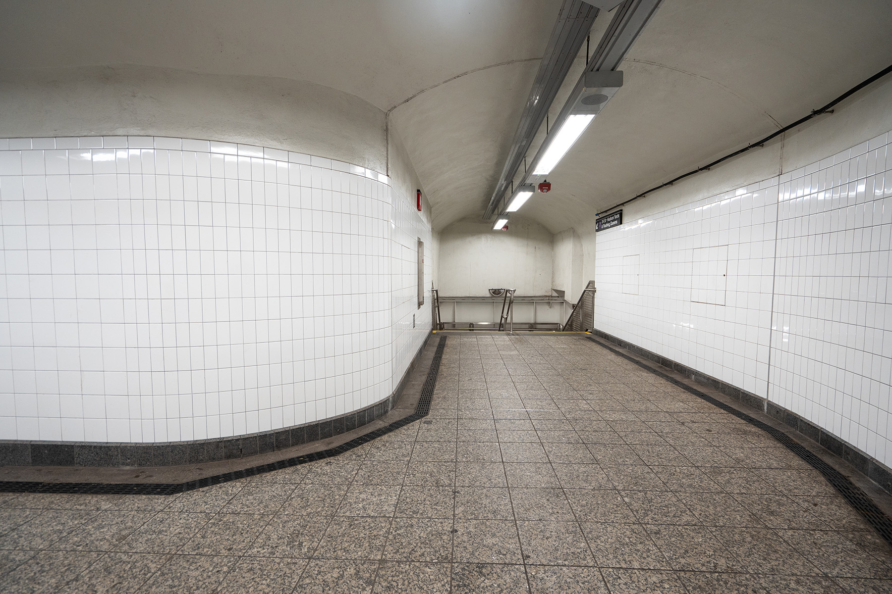 The completed tunnel has gray tile floors, white tile walls, and a white painted concrete ceiling. Long, thin rectangular-shaped lights line the hallway.