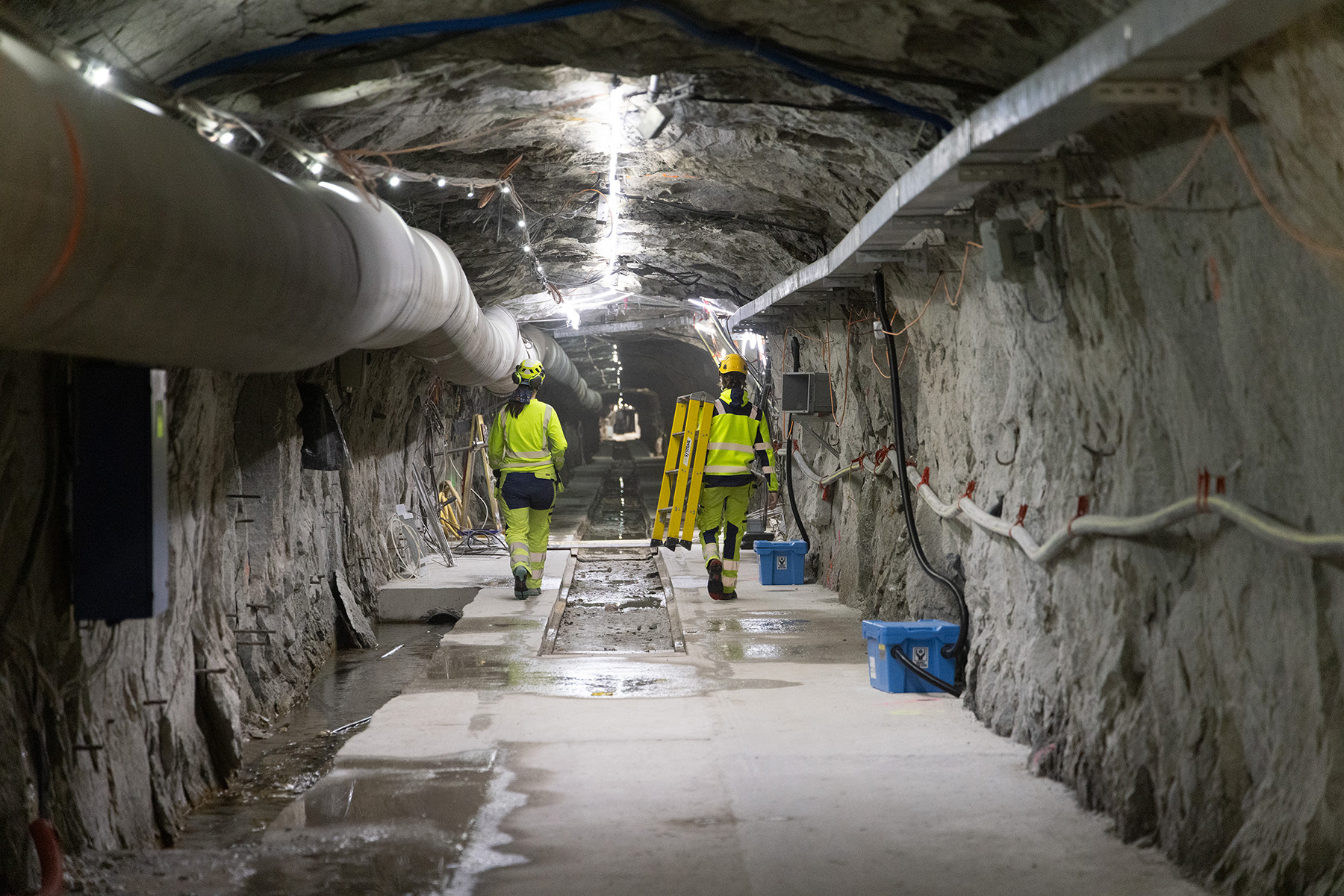 Two people wearing neon yellow long-sleeved shirts, pants, and hats walk along a sidewalk that lines both sides of a brightly lit underground tunnel. In the center of the tunnel is a long, narrow, rectangular-shaped opening in the ground. 
