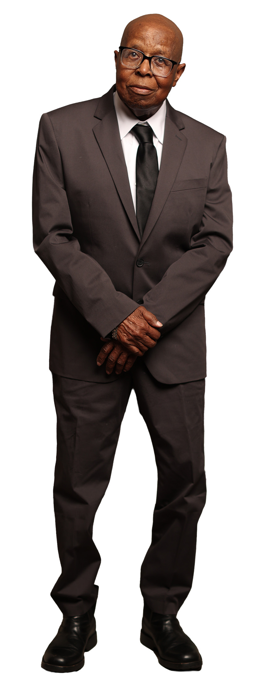 An older, bald man with brown skin wears a brown suit with a tie and glasses looks out at the camera. 