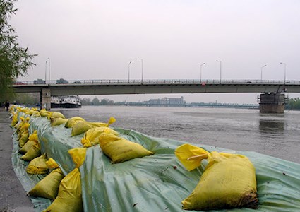 A row of large yellow sandbags flood barricade protecting river against flooding with bridge in background
