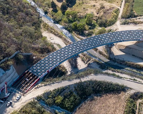 Aerial view of a large arched lattice bridge spanning a river with construction equipment near one bridge base.