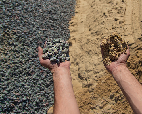 A pile of gravel on left and sand on right with a man’s hand holding a fistful of gravel in left hand and sand in the right