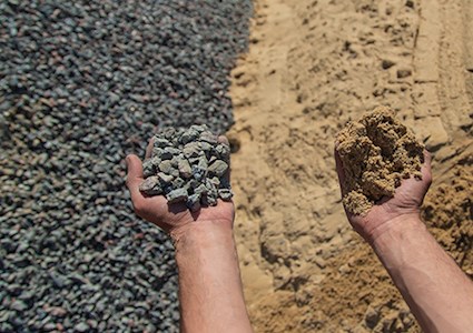 A pile of gravel on left and sand on right with a man’s hand holding a fistful of gravel in left hand and sand in the right